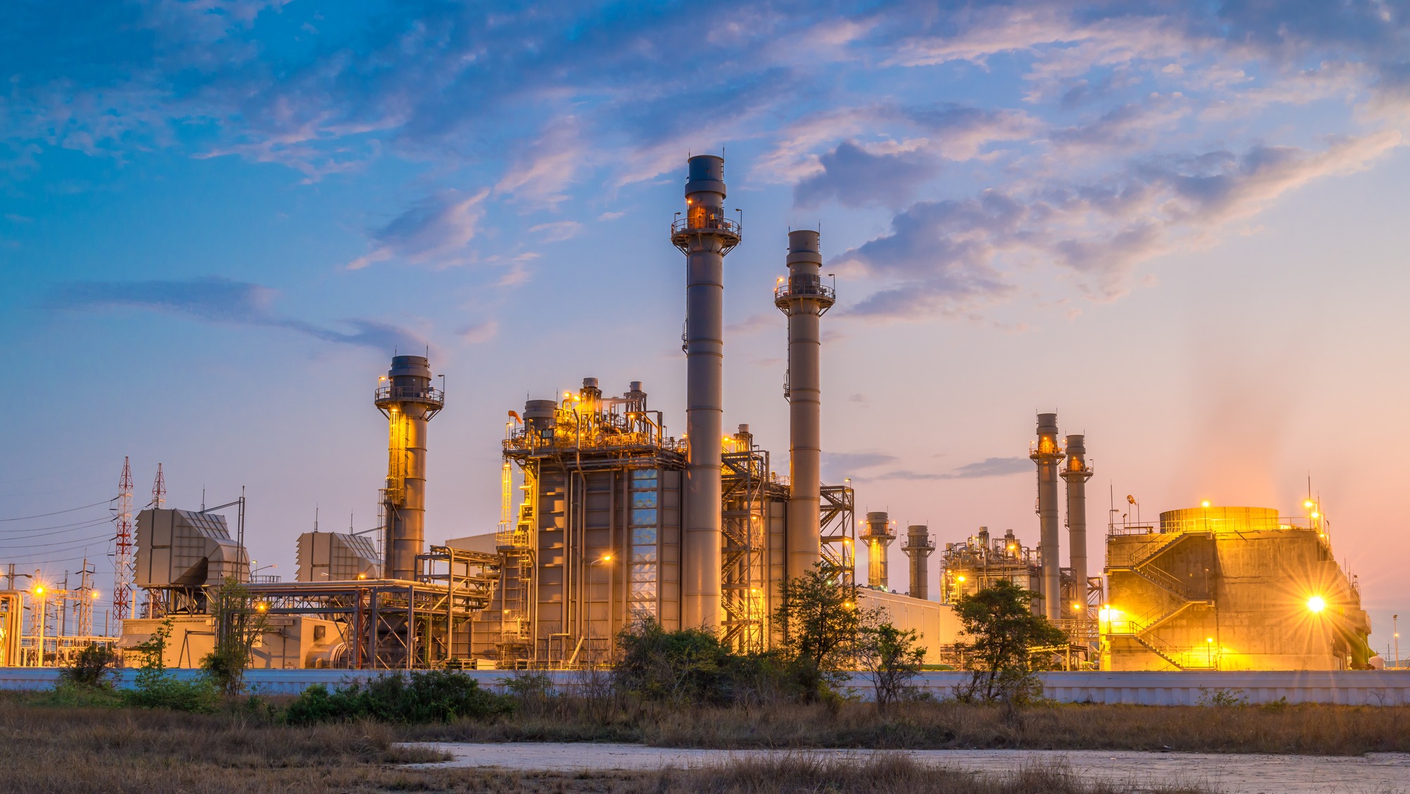 Gas-fired power plant illuminated at dusk, showcasing smokestacks and industrial structures, highlighting energy production infrastructure.